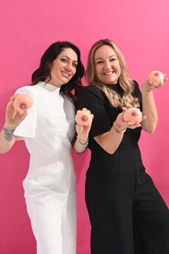 Gemma Bowers and Vikki Clark from The Tata Foundation stood together in front of a pink background smiling while holding two small silicone breasts each.