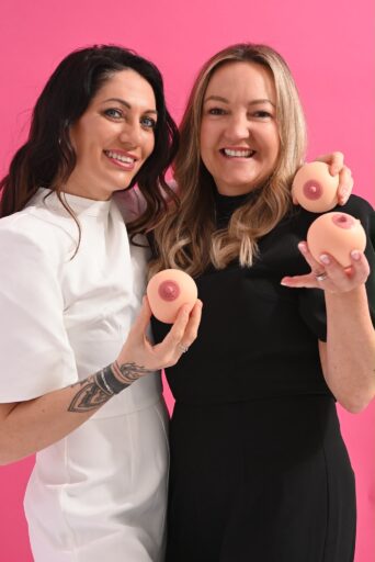 Gemma Bowers and Vikki Clark from The Tata Foundation stood together in front of a pink background smiling while holding two small silicone breasts each.